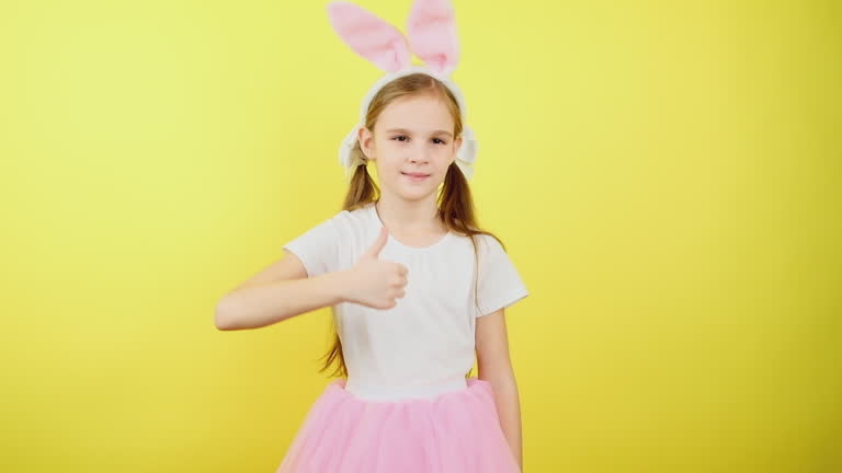 Portrait preteen girl in bunny fluffy ears doing winner gesture showing thumb up, isolated on yellow studio background. Easter holiday and emotion concept