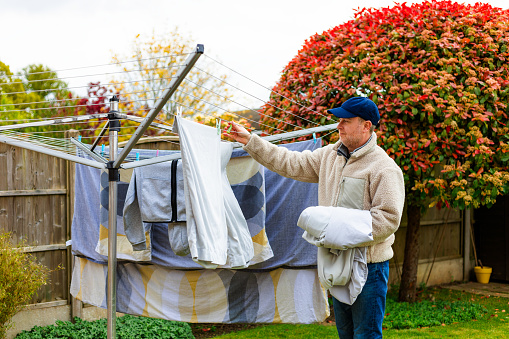 Portrait of a mid adult male in his 30s taking clean clothes and sheets from the clothesline in his garden at home. The man wears a beige fleece, navy blue baseball cap and blue denim jeans.