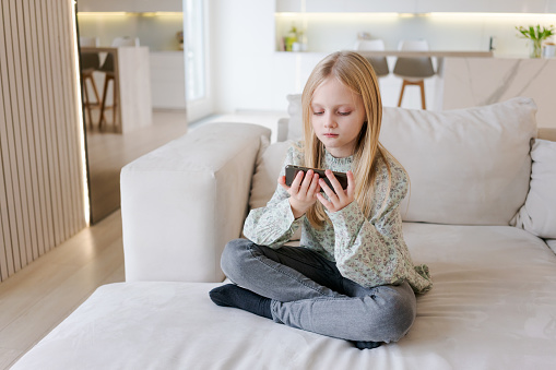 A young girl is sitting on couch and looking at her cell phone. Concept relaxation and leisure, as the girl is not engaged in any other activity