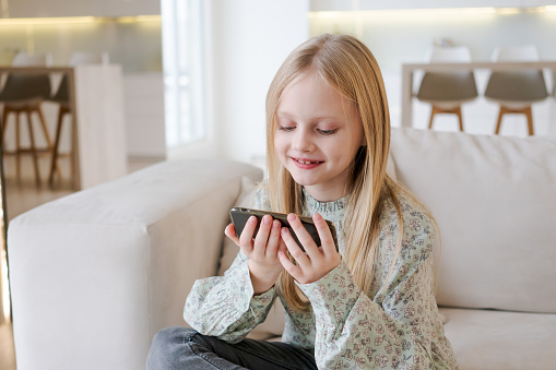 A young girl is sitting on couch and playing a video game on her phone. She is smiling and she is enjoying herself