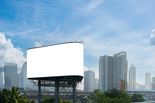 A large billboard is sitting in the middle of a city. The sky is clear and blue, and the city is bustling with activity. The billboard is white and blank, clipping path.