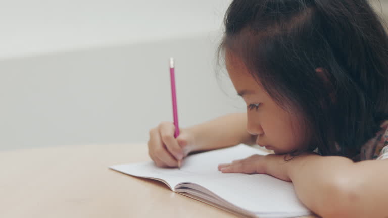 Face close up and selective focus of adorable Asian kid girl who is concentrating on writing on her notebook shows concept of education and studying of child in classroom for school learning.