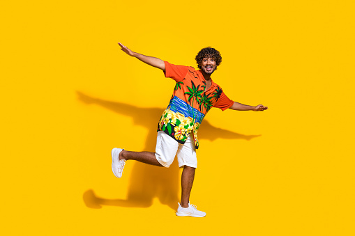 Full length photo of funky cheerful guy wear hawaii print shirt arms like wings fooling around isolated on yellow color background.