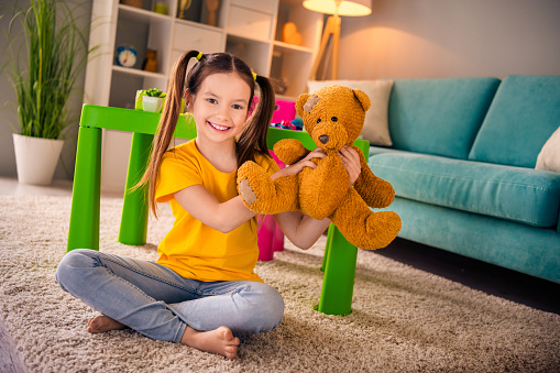 Photo portrait of adorable small girl sit floor hold show teddy bear toy dressed stylish yellow garment preschoolers kindergarten interior.