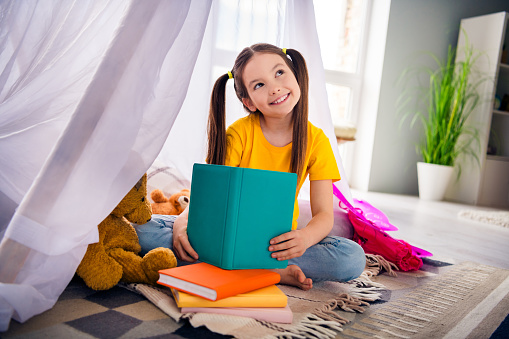 Photo portrait of adorable small girl read book tent look empty space dressed stylish yellow garment preschoolers kindergarten interior.