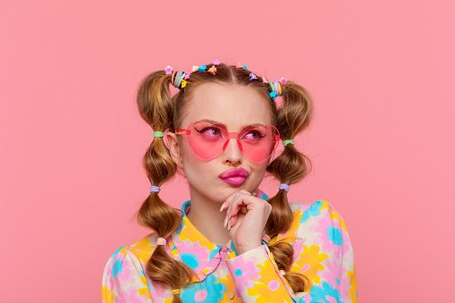 Confused gen Z teen girl with stylish hairstyle with hairpins wearing colorful floral pattern shirt and pink eyeglasses looking away with hand on chin. Studio shot, pink background.