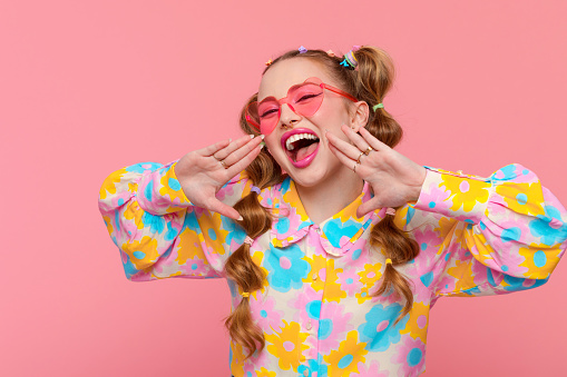 Excited gen Z teen girl with stylish hairstyle with hairpins wearing colorful floral pattern shirt and pink eyeglasses looking at camera and shouting. Studio shot, pink background.