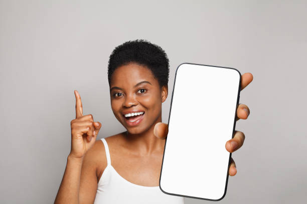 istock Pointing up healthy woman holding smartphone with white empty blank screen display on gray studio wall background. Happy model with phone 2149680914