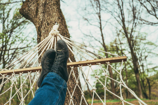 Man laying down and relaxing in back yard hammock attached to a tree, wearing jeans and dirty black sneakers, selective focus