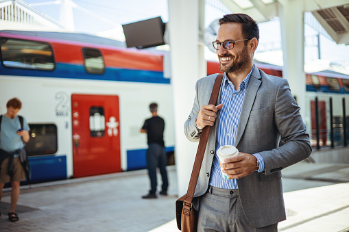 A traveling male business person is at the train station with a reusable travel mug of coffee. A man in a train station commuting to work. Businessman getting in passenger train and going home after working day.