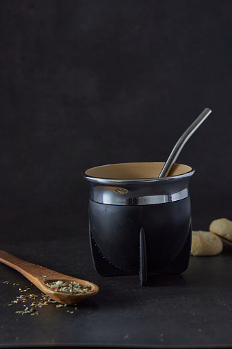 Yerba mate herb and calabash gourd with steel bombilla with a wooden spoon an two cookies on black background