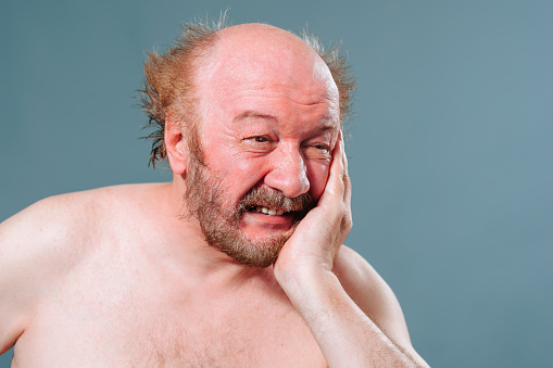 Toothache does not give rest, an undressed elderly man holds his cheek with his hand, a tooth is inflamed, a nerve hurts. Studio isolated blue background. Front view in studio.