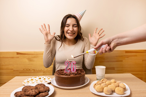A girl sits in front of a table with a festive cake, on which a candle is lit in the form of the number 26. The concept of a birthday celebration.