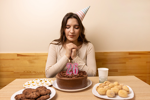 A girl sits in front of a table with a festive cake, on which she blows out a candle in the form of the number 26. The concept of a birthday celebration.