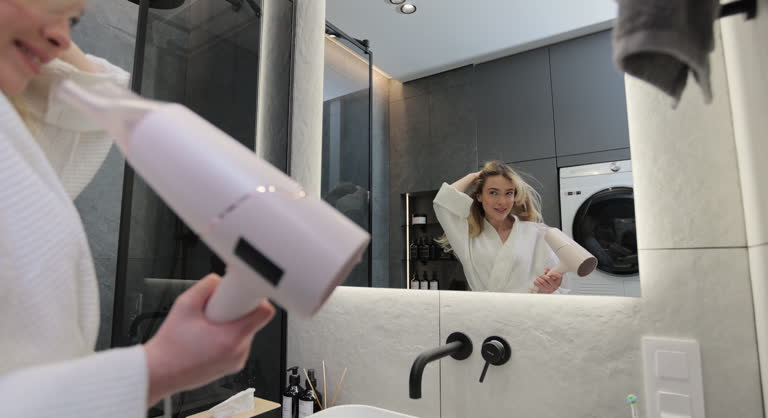 Young woman in bathrobe drying hair with a hairdryer in the bathroom at home. Usual morning beauty treatments in the bathroom.