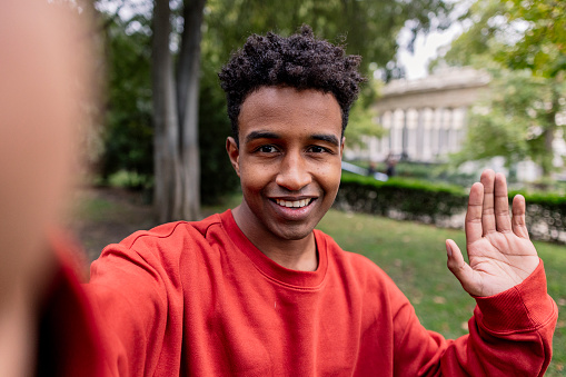 A charming young man with a friendly smile warmly greets the camera with a wave, set against a verdant park backdrop.