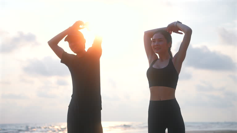 Happy asian sports couple stretching exercises at seashore with dramatic sky and bright sunset. Silhouette of man and woman jogger during outdoor activity on beach. 4k resolution and slow motion shot.
