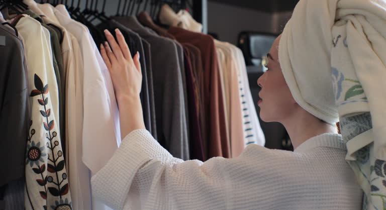 Woman wearing a bathrobe and a towel on her head choosing clothes from rack with hangers inside room. Lady looks to clothes, decides what to wear. Lifestyle women relax at home concept.