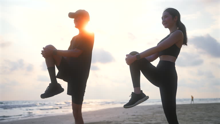 Happy asian sports couple stretching exercises at seashore with dramatic sky and bright sunset. Silhouette of man and woman jogger during outdoor activity on beach. 4k resolution and slow motion shot.