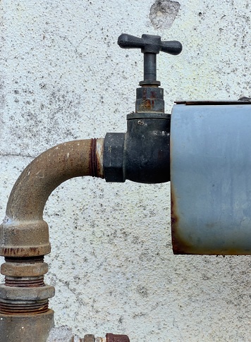 A close-up captures the rustic charm of an old metal water tap with a classic round wheel handle, affixed to a corroded and bent pipe. Set against a backdrop of a weathered concrete wall.