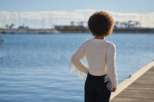 A woman stands on a dock overlooking a body of water. She is wearing a white sweater with fringe and black pants. The scene is peaceful and serene, with the water reflecting the sky