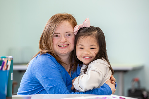 Young woman with Down Syndrome smiles and embraces elementary age girl who also has Down Syndrome