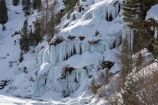The stunning scene of a frozen waterfall is lit up by the gentle sunlight, making a beautiful mix of light and shadows. Huge icicles hang from the cliff, and the slopes covered in snow with evergreen trees add to this peaceful mountain picture. A natural glow from the sunlight adds a magical touch, perfectly capturing the calm and impressive feeling of winter in the mountains.