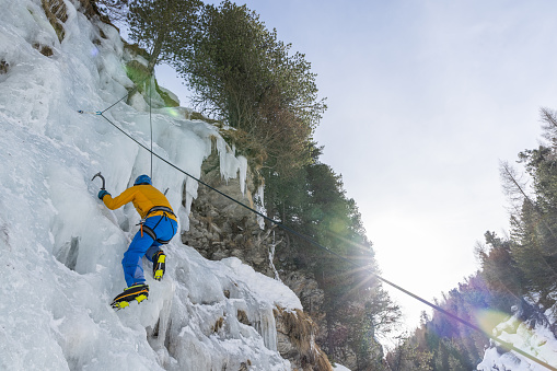 An ice climber in bright yellow and blue climbs a frozen cliff, standing out against the ice and blending with sunlight coming through the trees. The picture shows the climber's skill and the excitement of ice climbing, set in a stunning winter landscape where the ice's power meets the beautiful sunlight.