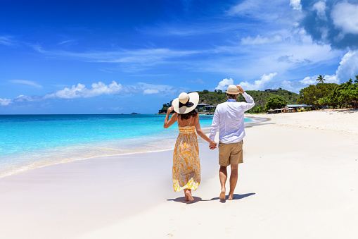 Happy holiday couple with sunhats walks down a tropical beach with turquoise sea in the Caribbean, Antigua island