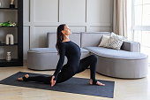 Woman practicing yoga in the room, performing a variation of Anjenyasana exercise, half moon pose, working out in black sportswear on a mat, full length view