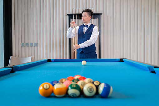 A sharply dressed man in a vest and bow tie focuses intently while lining up a shot on the blue felt of a billiard table.