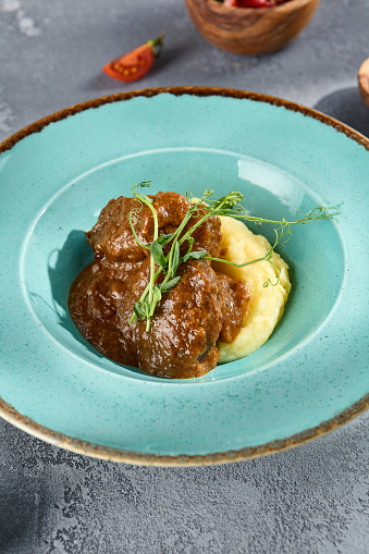 Braised beef cheeks on mashed potatoes in a rustic setting with tomatoes and peppercorn on textured grey background.