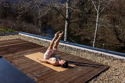 Mid adult woman doing yoga, exercising, stretching and relaxing on rooftop deck surrounded with nature at spring. She is wearing pajamas and bikini top and she is doing her morning exercise and enjoying sunlight