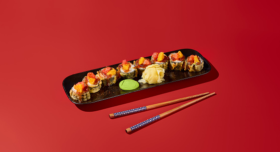 Tempura sushi roll with tangy citrus sauce, served on a black plate. Modern still life on a deep red background with hard shadows. Ideal banner for an Asian restaurant.