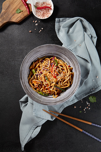 A vertical top view of a luscious bowl of Udon noodles mixed with chicken and vegetables. The garlic sauce, chopsticks, and textile on the black background enhance the visual depth of this Asian culinary delight.