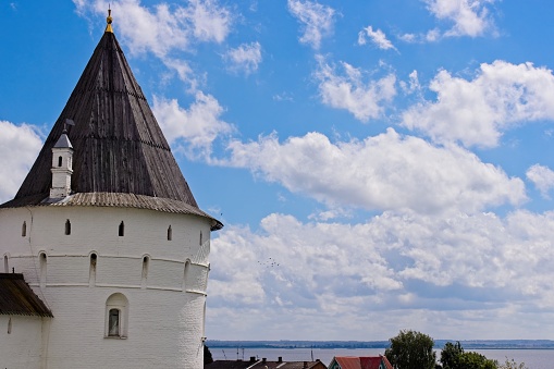 Magnificent view of a round watchtower with a hipped wooden roof and narrow loopholes, white stone walls, fabulous view, medieval Russian architecture, city sights, distant view of the lake, sky and clouds, cityscape, summer.