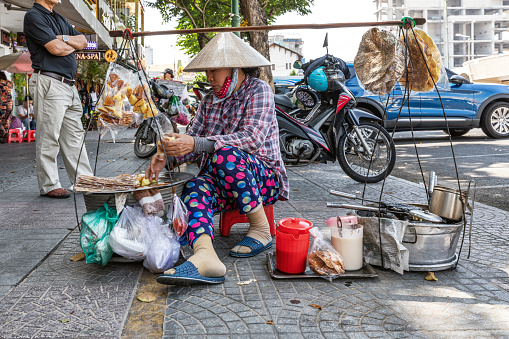 Ho Chi Minh, Vietnam - March 12, 2024: Woman in a traditional conical hat, sells snacks such as poppadoms and cakes on a street in the center of Ho Chi Minh.