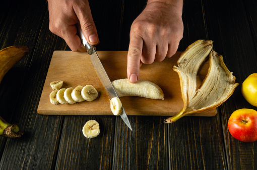 Chefs cutting banana on a wooden boards. Low key concept of preparing a fruity breakfast dish on the kitchen table.