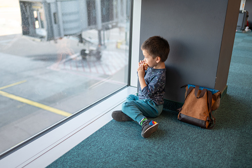 A toddler boy, around 3 years old, sits by an airport window eating a snack as he waits for his flight, capturing the essence of travel with young children. A curious toddler indulges in a tasty snack while seated by the airport window looking at the airplanes taking off and landing, captivated by the bustling activity outside during their travel adventure