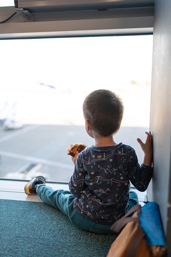 Back view of a toddler boy, around 3 years old, sits by an airport window eating a snack as he waits for his flight, capturing the essence of travel with young children. A curious toddler indulges in a tasty snack while seated by the airport window looking at the airplanes taking off and landing, captivated by the bustling activity outside during their travel adventure