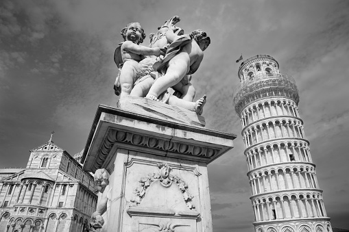 Pisa, Italy - 2010, October 30 : A statue of the 19th century Putti Fountain in front of the leaning tower and the cathedral of Pisa in Tuscany, Italy  - monochrome