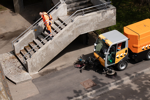 Basel, Switzerland - April 18, 2024: Worker wearing orange uniform cleaning city bridge stairs with leaf blower and a street sweepers vehicle cleaning the sidewalk
