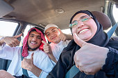 An Arabic family, dressed in traditional attire, smiles happily in a car, showing thumbs-up signs. They seem excited for a fun picnic adventure together