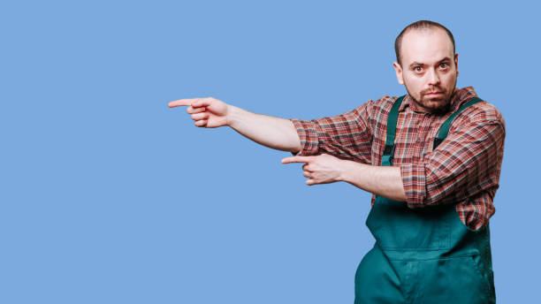 istock Young and attractive man, dressed in a coverall, with a quizzical expression as he points 2149721695