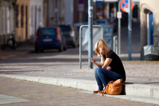 Aalborg, Denmark. October 1, 2011. Young blond woman with head in hands messaging online over smart phone while sitting by empty street on footpath in city during sunny day