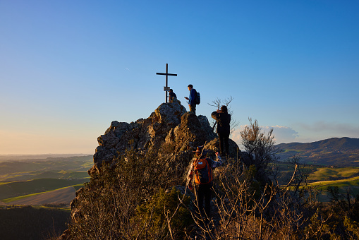Volterra, Italy. March 12, 2023. Active male hikers climbing on top of rocky mountain towards wooden cross against clear blue sky with beautiful landscape in background during sunset