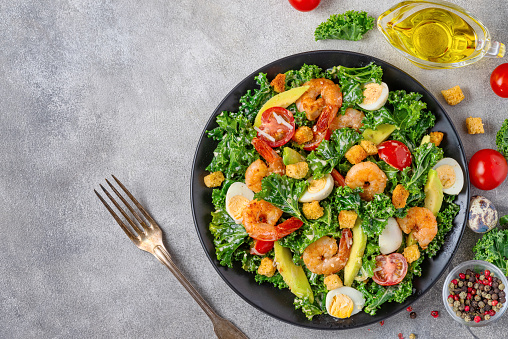 Delicious seafood salad with shrimps, kale cabbage, avocado, tomatoes and quail eggs in a plate with fork on gray concrete background. Top view with copy space. Healthy diet food for dinner.