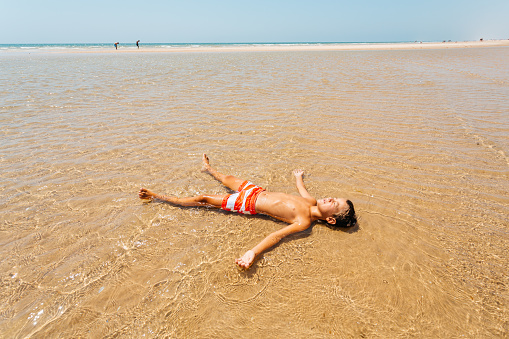 Boy lying in the shallow water and enjoys the sun, sunbathing on his summer vacation in the Algarve region in Portugal