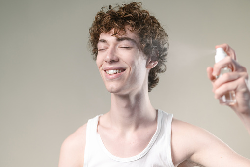 A young man in a white undershirt spraying his face and smiling