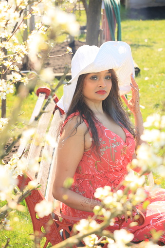 A Fijian model sitting outside on a bench on a sunny Spring day with flowering tree and green background. She is wearing a big white hat, long, black hair, makeup, and a red sleeveless dress.